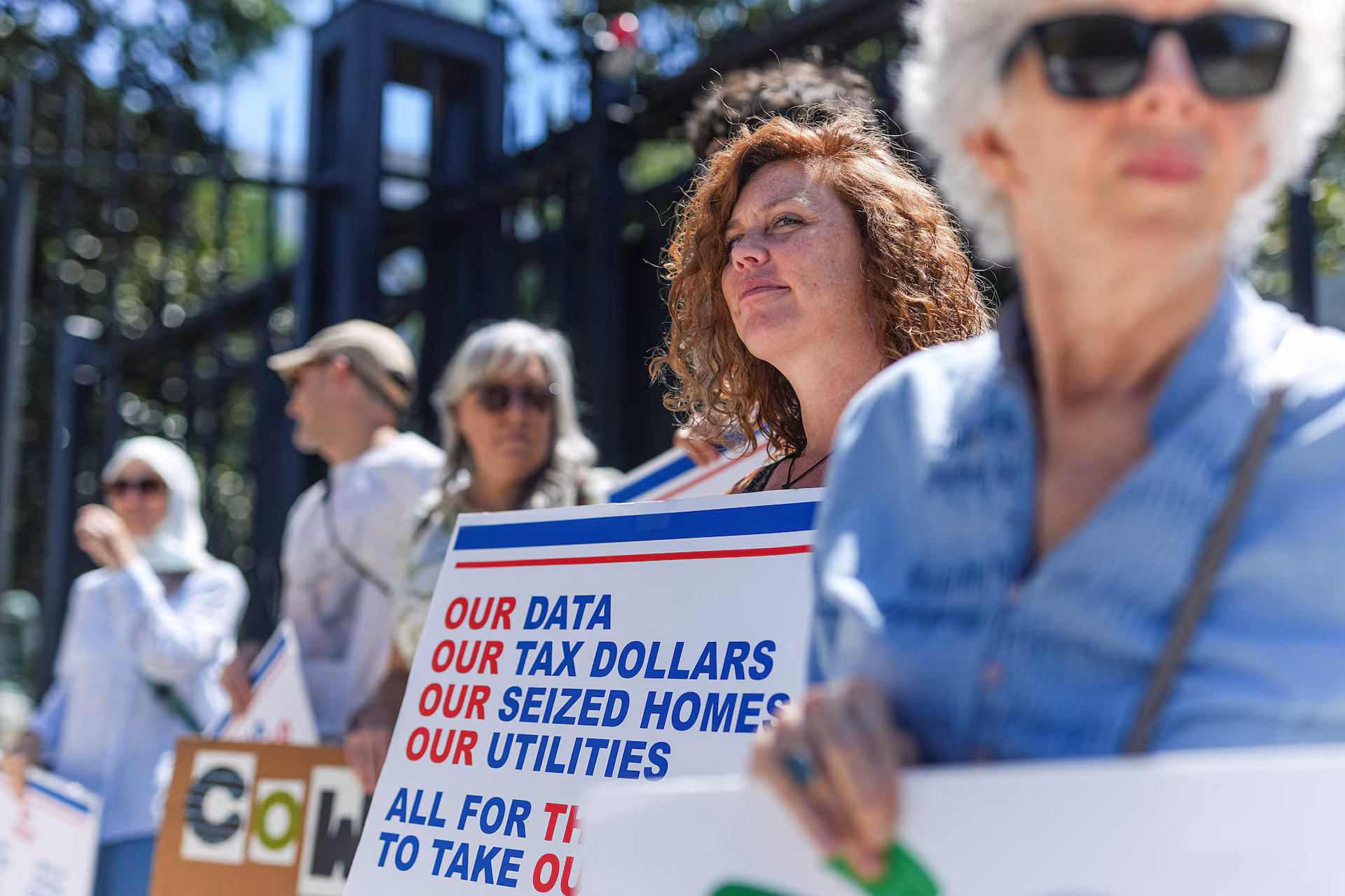 Protesters outside a fenced site hold a sign reading Our Data, Our Tax Dollars, Our Seized Homes, Our Utilities — All For Them To Take