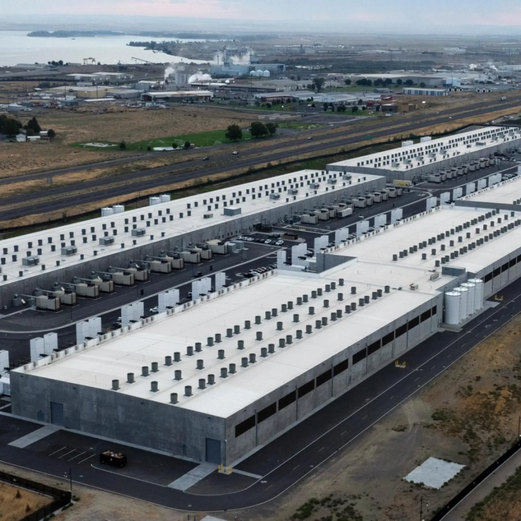 Aerial view of a sprawling data center campus of long gray warehouse buildings beside a highway, with farmland and a river in the distance